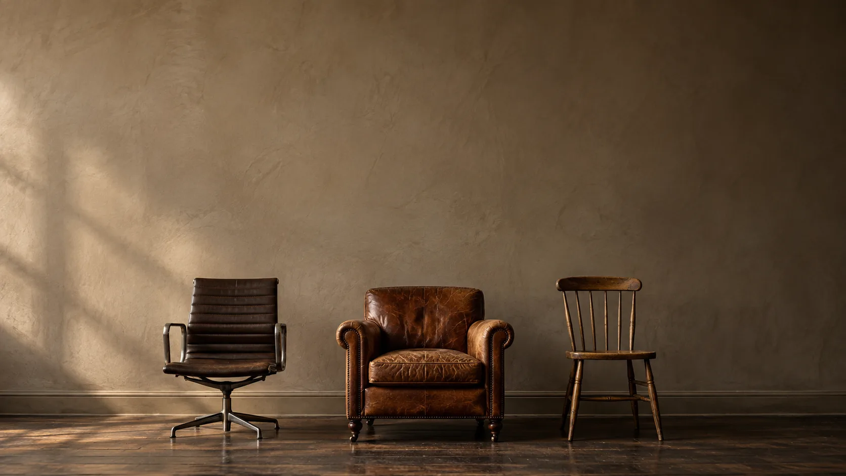 Three different chairs side by side against a plain plaster wall: an office chair, a leather armchair and a wooden chair.