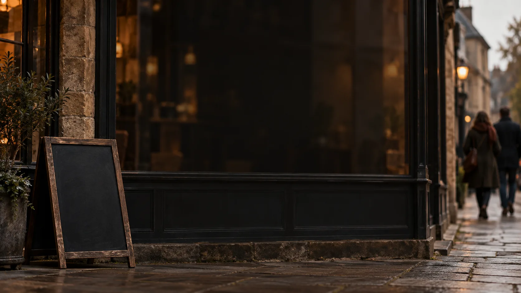 A shop window in a market town with a blank chalkboard outside and two passers-by on the pavement.