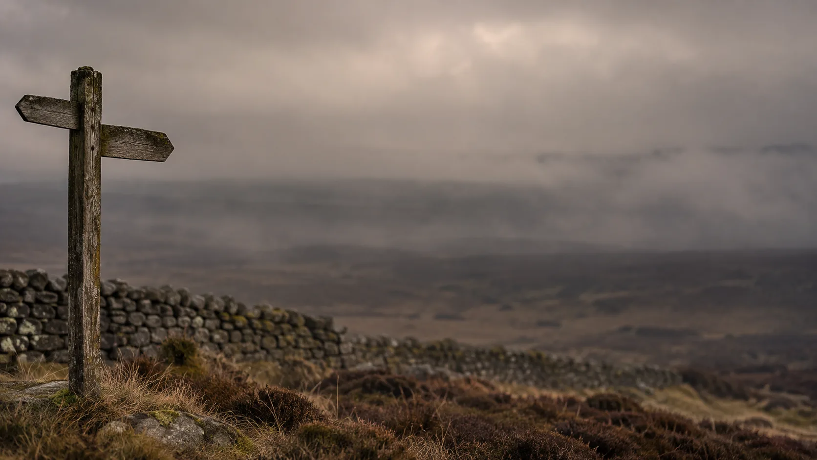 A weathered wooden signpost with blank arms on a misty moor, a dry-stone wall running away behind it.